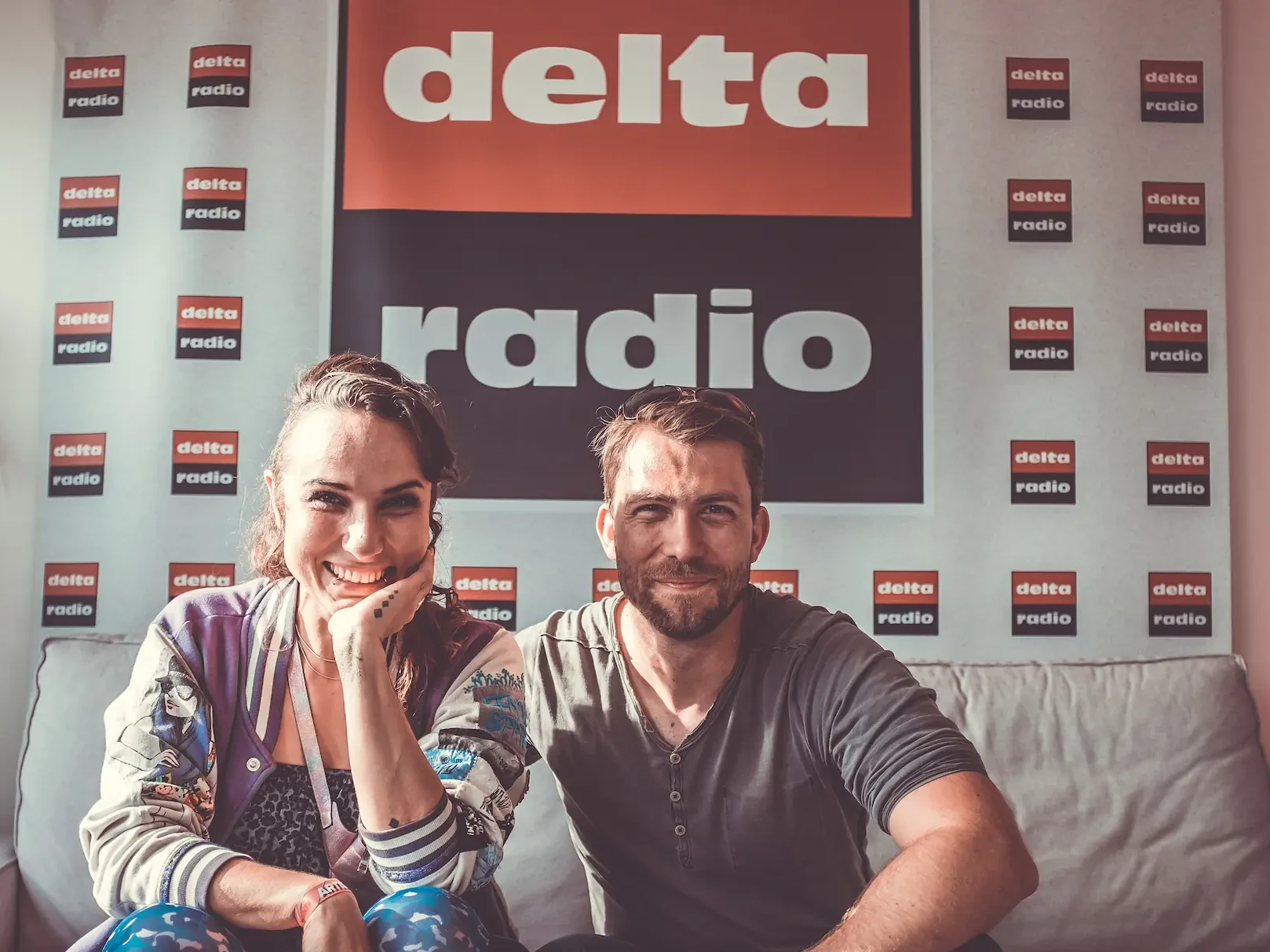 delta radio Frau und Mann sitzen lächelnd auf der Couch vor einer Logowand von delta radio.